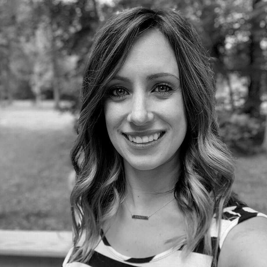 Portrait of Kristy Wilkinson smiling warmly at the camera, with shoulder-length wavy hair, wearing a striped sleeveless top and a delicate bar necklace, outdoors with blurred greenery in the background, in black and white