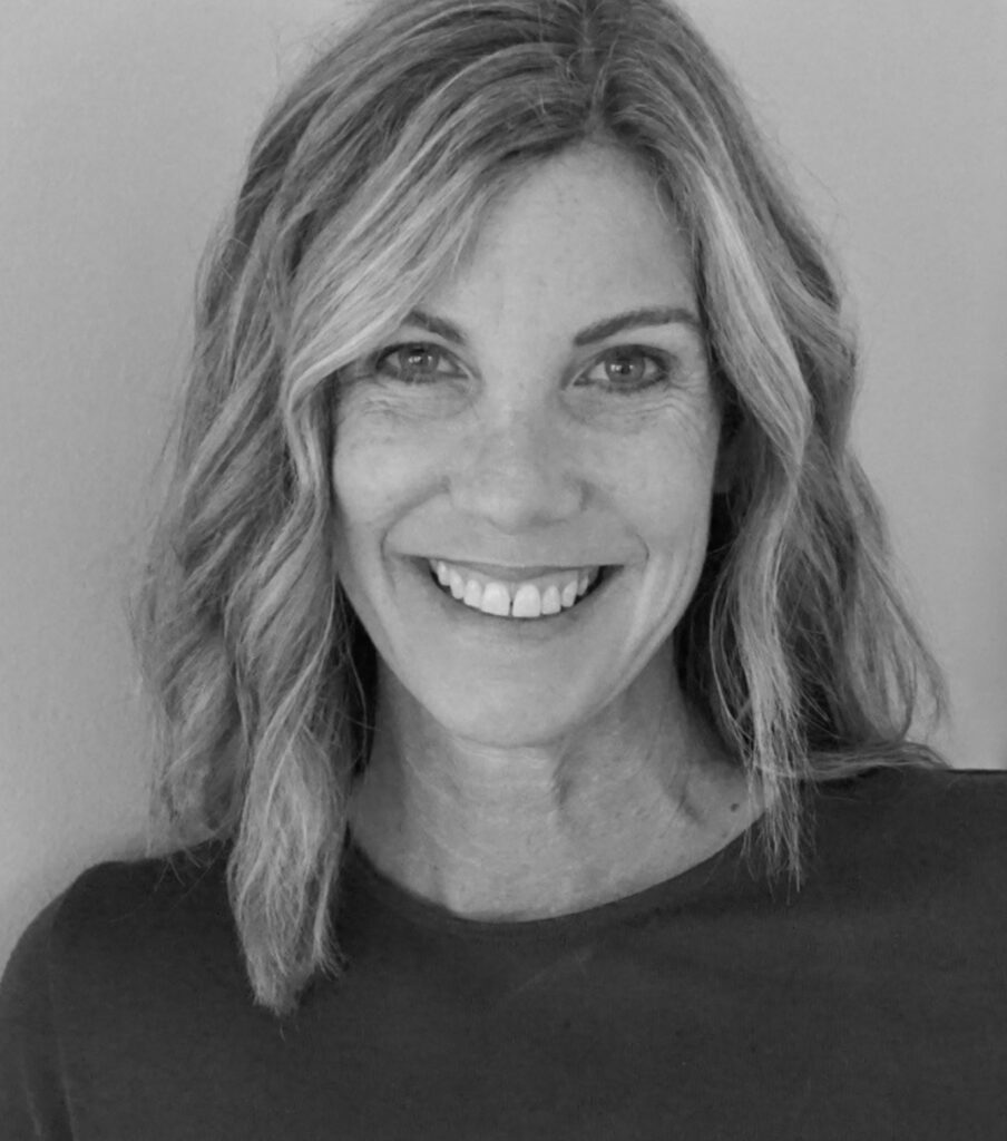Professional headshot of Laura Goeglein, a woman with shoulder-length wavy hair featuring lighter highlights, smiling warmly at the camera while wearing a dark crew-neck top against a plain light background, in black and white